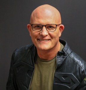 smiling man with bald head wearing square frame glasses. He is clean shaven, wearing an army green jacket underneath a black leather jacket with the superman lol embossed into the leather.