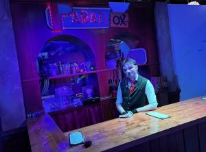 Semi-masked bartender standing behind a strange bar named, Paradox.