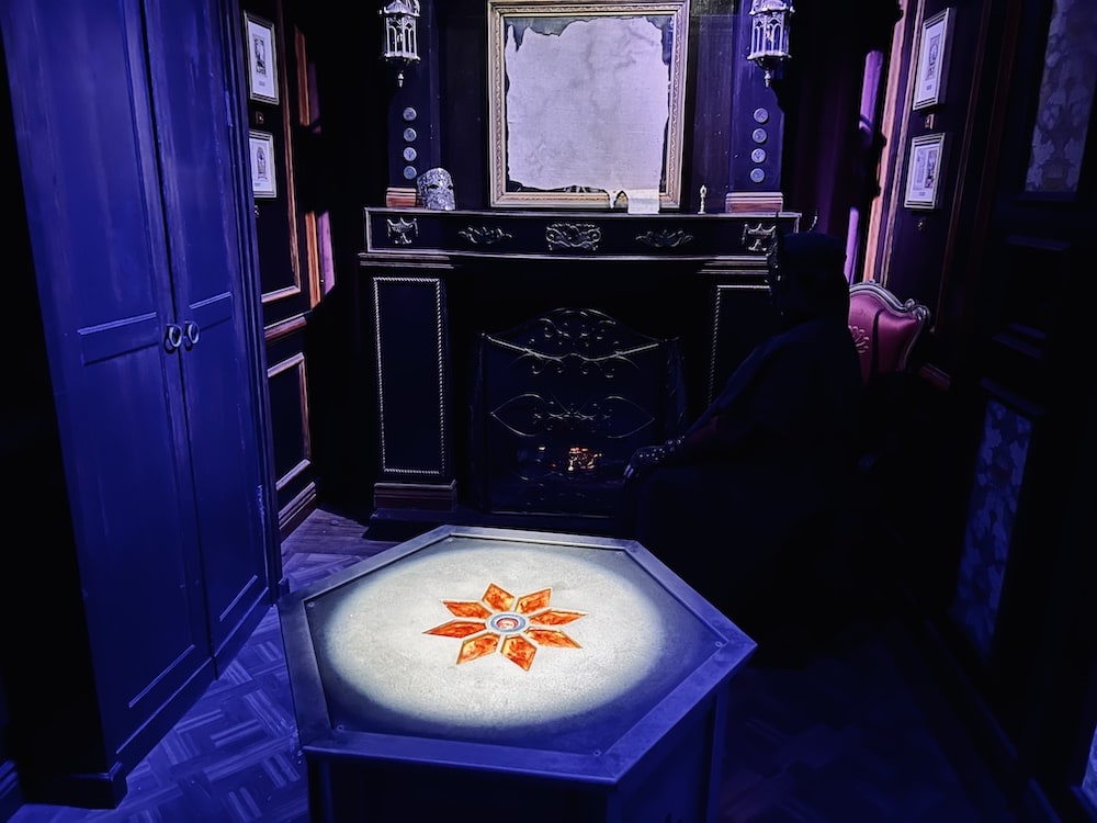 An illuminated pedestal with an orange sun inlay in the middle of a victorian study. A cloaked and masked figure sits beside a smoldering fireplace.