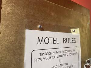 A sign mounted in clear acrylic reads, "Motel Rules: tip room service according to how much you want them to forget."