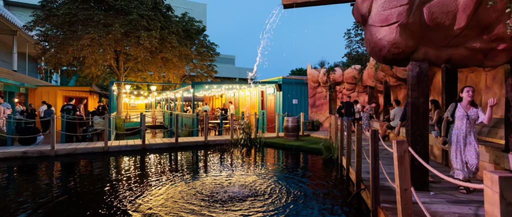 outdoor area at phantom peak. there is water trickling down into a pond. surrounding the pond is a walkway filled with people. in the distance we can see brightly lit bars and shops.