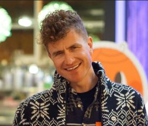 smiling man with curly hair short on the sides and longer on top, in a patterned black and white jacket.