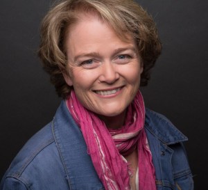 the image is a headshot of a mature, smiling woman with short blond hair and blue eyes in a pink scarf and denim jacket.