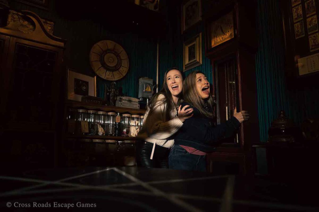 Two women cowering in the corner clutching a grandfather clock in a dark room.