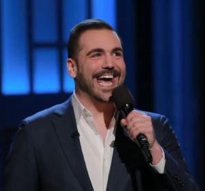 Image of a man in a dark blue suit and white button down shirt. He has neat, short brown hair and a short beard. He's holding a microphone with a smiling, excited expression.