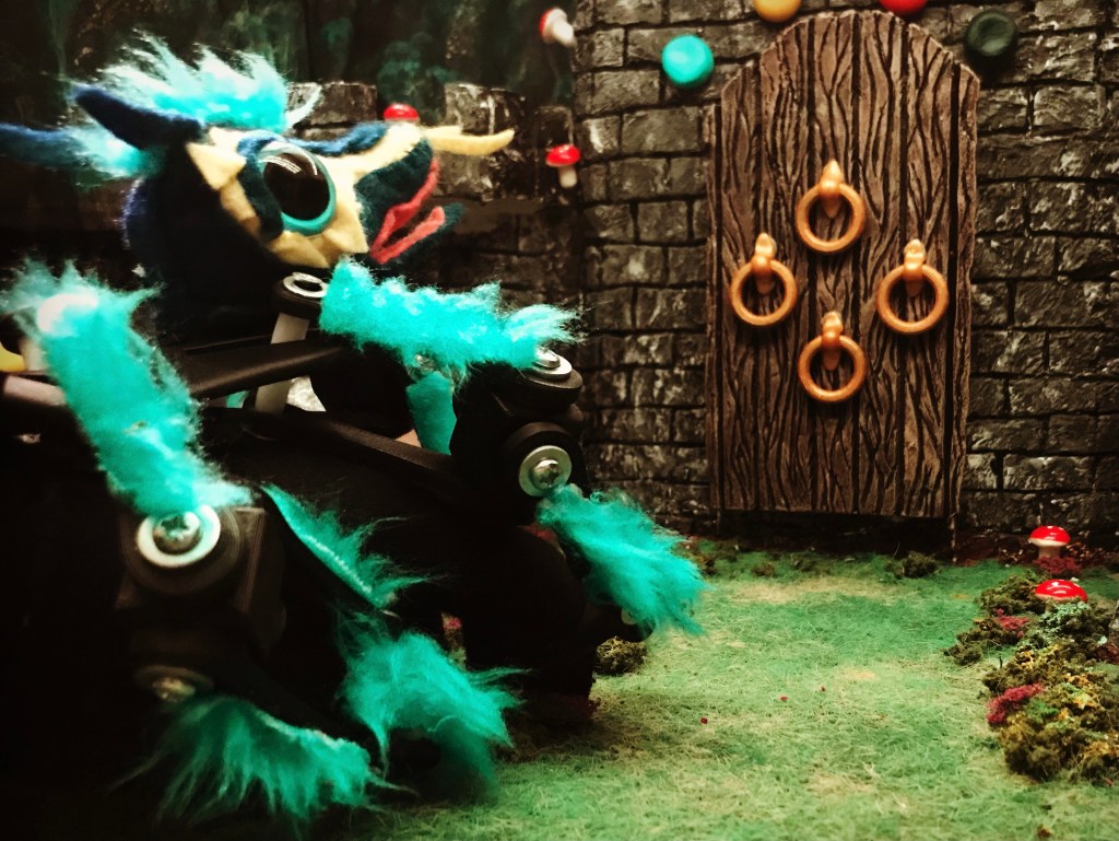 image of the "keeper". it is a hand puppet of a spider dragon with large cute eyes and teal fur. it's facing a miniature door decorated with 4 knockers.