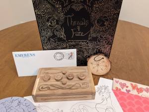 Black Threads of Fate box with an intricate silver pattern printed on it. Beside the box are a few puzzle components including a beautiful puzzle box and a wooden medallion with runes etched into it.