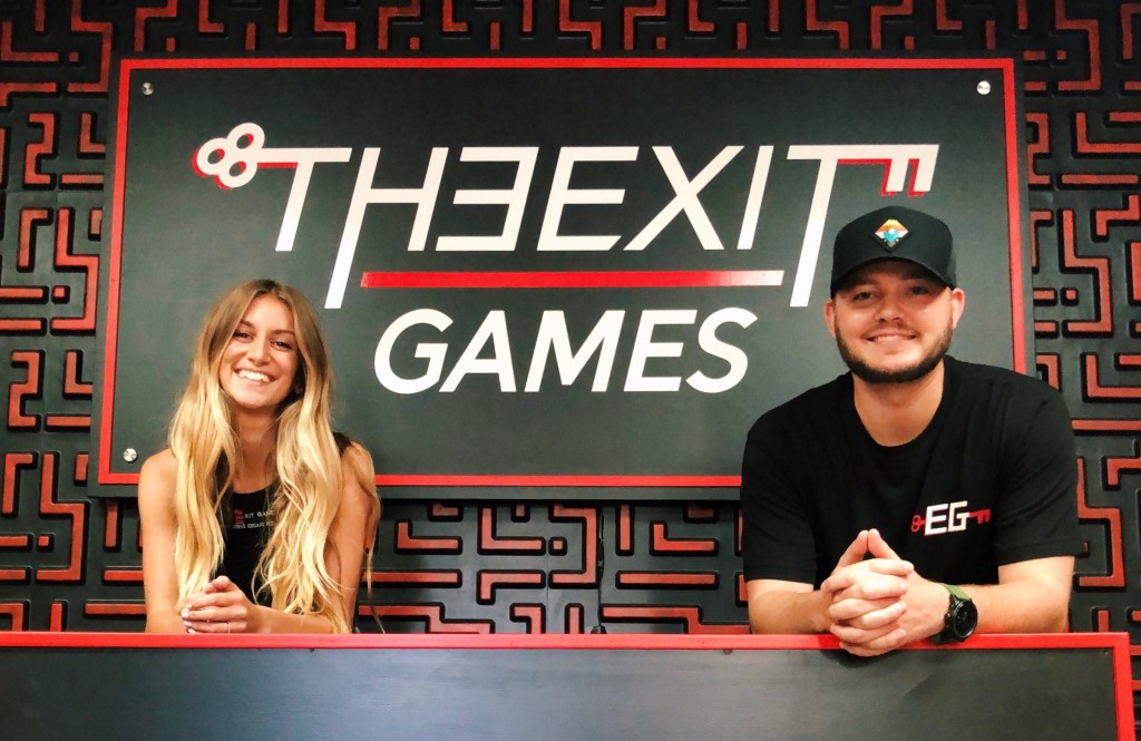 A man and a woman leaning on a counter in front of a large sign that says "The Exit Games". The woman is smiling with long wavy blond hair, wearing a black tank top. The man is smiling, with a short beard. His is wearing a black baseball cap and a black t-shirt with The Exit Games logo.