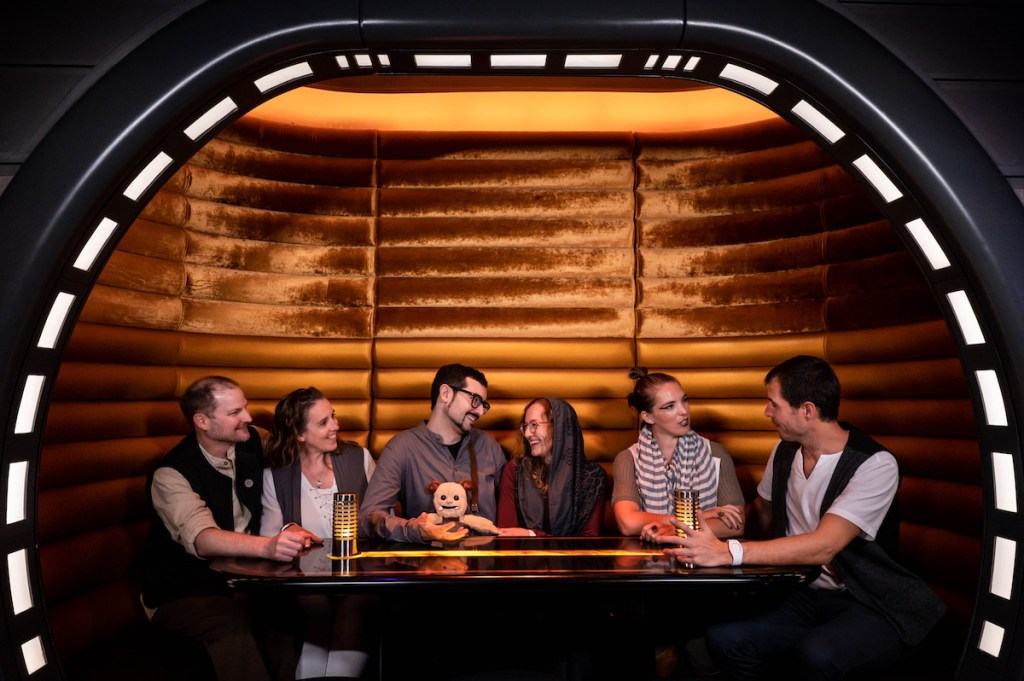 Richard, Heather, David, Moss, Lisa, Theresa, and Brendand talking at a table in the Sublight Lounge of Star Wars Galactic Starcrisier.