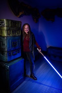 Lisa emerging from behind a stack of crates holding a lightsaber downwards.