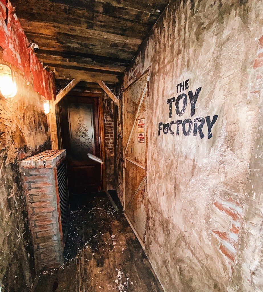 A long, rundown corridor. "The Toy Factory" is painted on the wall, the "a" in factory has been replaced with the face of a Teddy Bear.