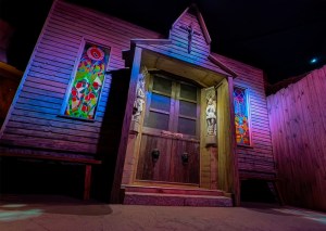 Exterior of a church at night. It has elaborate statues besides its doors, and a pair of stained glass windows on either side, and a large crucifix above the door.
