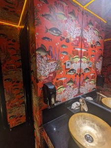 A bathroom with brass fixtures and coral reef wallpaper. The ceiling is outlined in yellow LED strips.