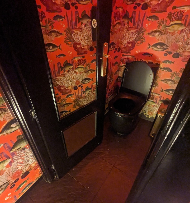 A black toilet surrounded by walls clad in coral reef wallpaper.