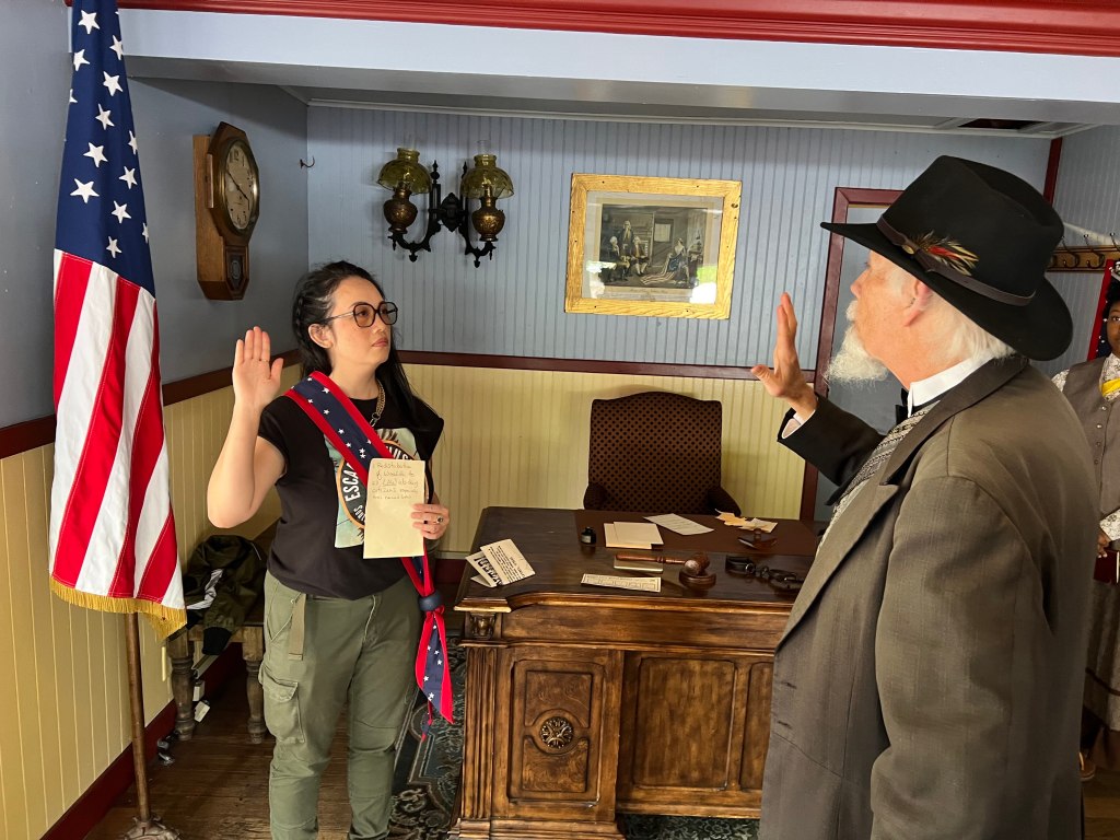 An asian woman wearing a mayoral sash with her hand up being sworn in by an older man in a cowboy hat and old fashioned suit. They are in a mayor's office with an american flag and a desk.