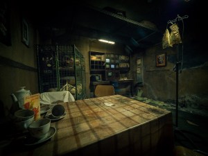 A rundown room in a hospital, a table and chairs sit in the center with empty teacups atop. Beside the table is an IV bag holder, in the distance is assorted equipment.