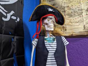 A skeleton wearing a pirate costume and sitting beside a map and a pirate flag.