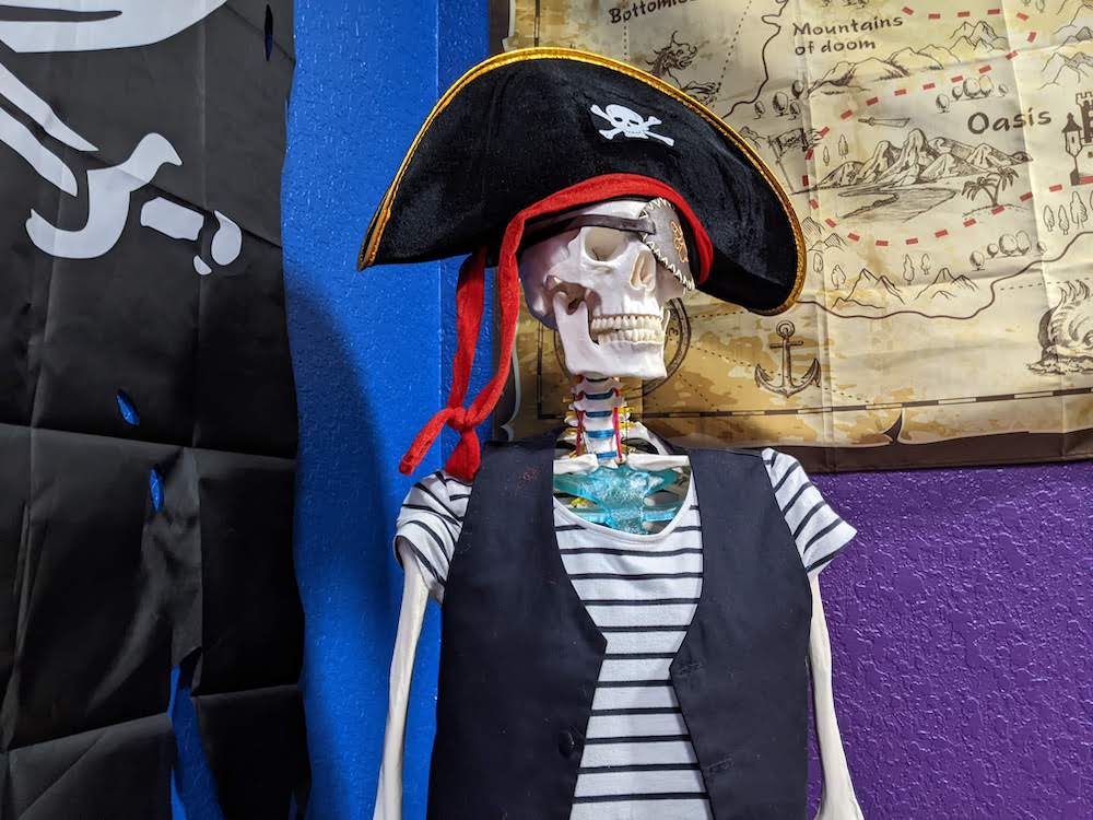 A skeleton wearing a pirate costume and sitting beside a map and a pirate flag.
