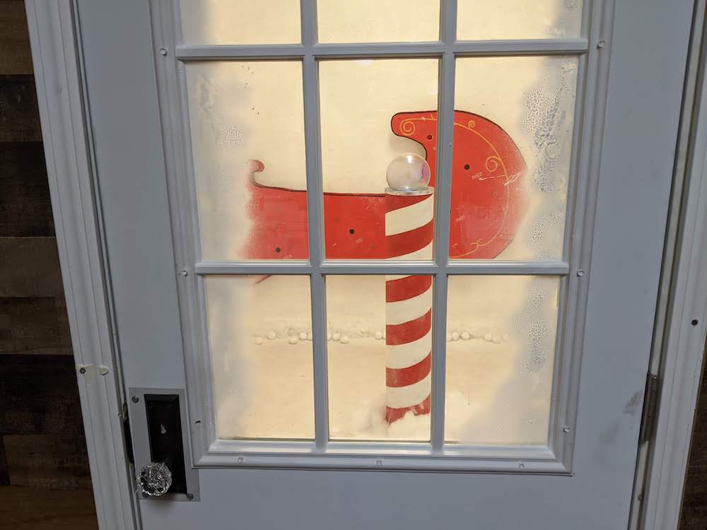 Closeup of a door house, through a window you can see Santa's sleigh and a pole painted red and white like a candy cane.