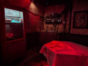 A dim, red lit room, with black netting, pumpkins, jack-o-lanterns, and a coffin covered in a sheet.