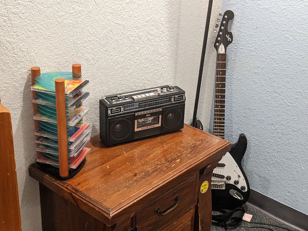 A guitar leaning against the wall beside a small boom box and a CD rack.