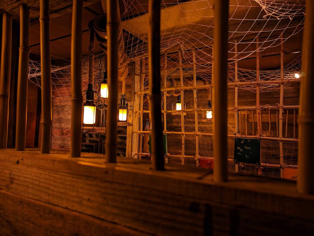 A dramatically lit wooden jail cell with white netting hung from the ceiling.
