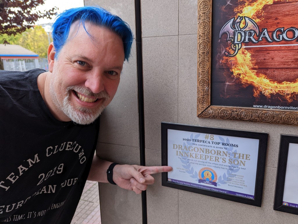 Man with blue hair and a grey beard wearing a black t-shirt, smiling while pointing to a framed award that says "2020 TERPECA TOP ROOMS: Dragonborn: The Innkeeper's Son"