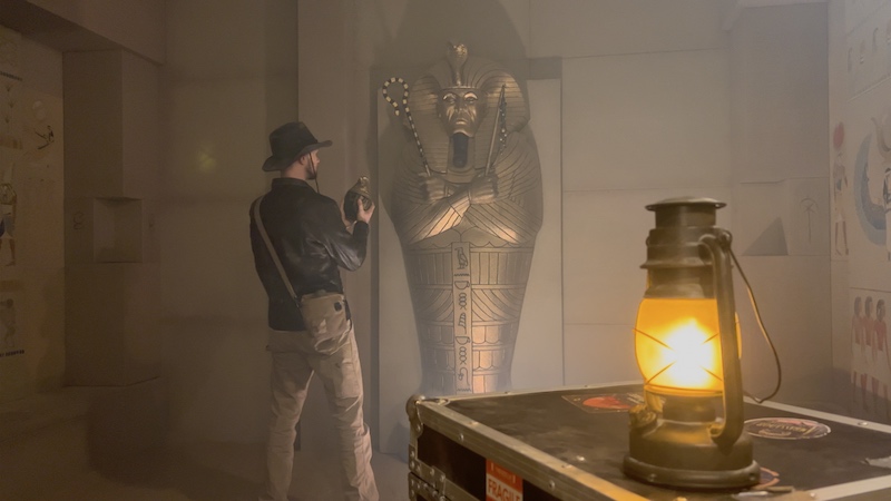 A man dressed as Indiana Jones in the middle of an Egyptian tomb set, standing in front of a sarcophagus with a canopic jar in his hand.