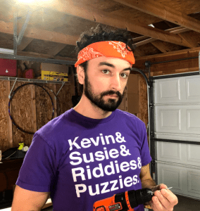 man looking serious in a garage holding an electric drill. wearing a purple t-shirt that says "keving & susie & riddies & puzzies" he has curly dark hair held back by an orange bandana and a short beard.