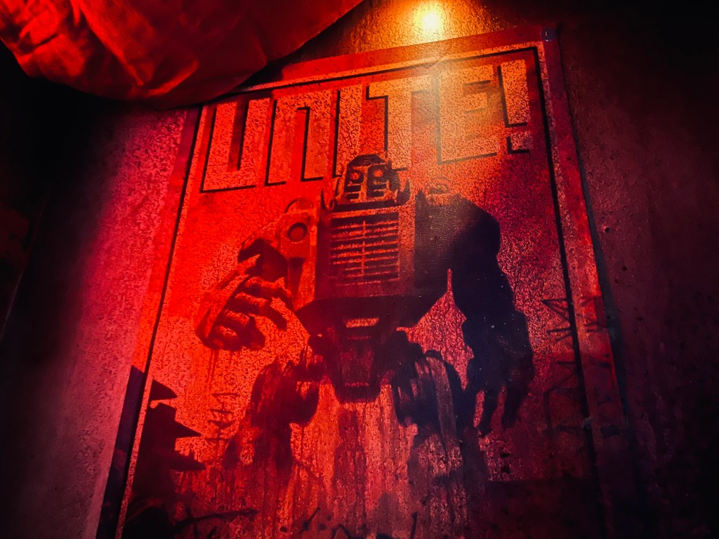 A red propoganda poster with an imposing robot on it. One word appears on the poster, "UNITE!"