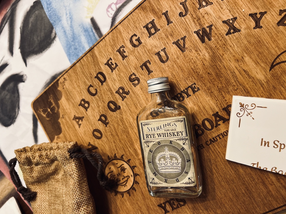 Closeup of a beautiful wooden spirit board, a small bottle of whiskey, a broken tile shard, a burlap bag, and some children's illustrations of ghosts.