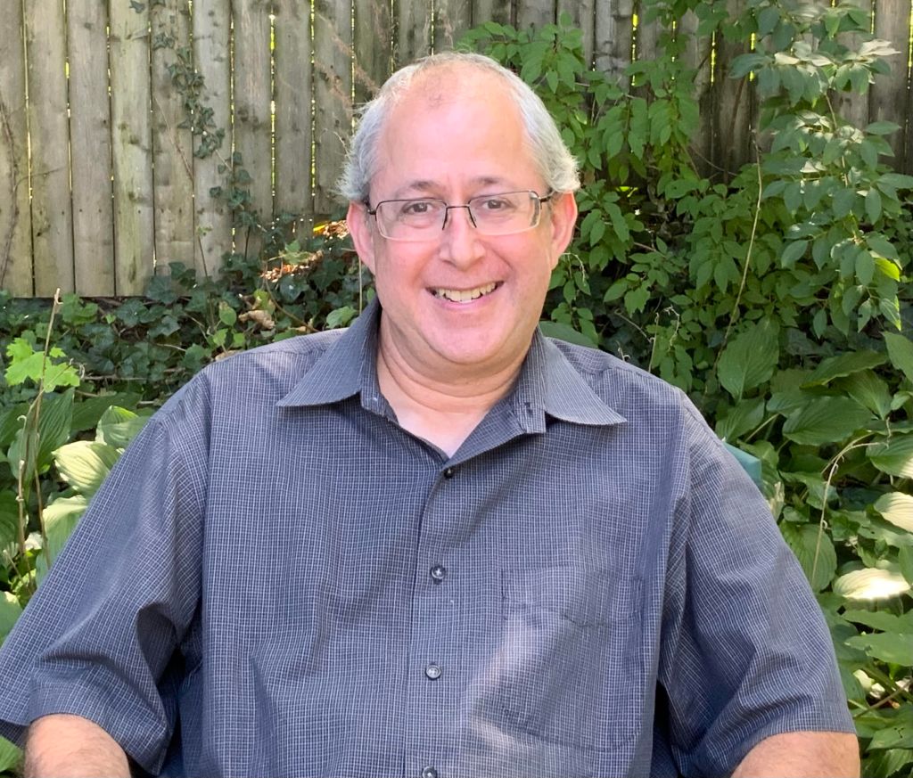 older man with white hair and glasses, smiling wearing a plaid blue button down shirt in a garden.