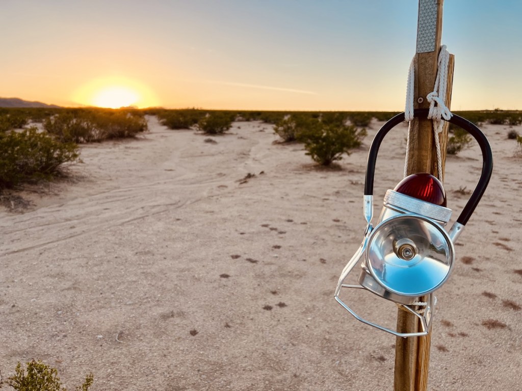 A lantern hung from a post in the middle of the desert. The sun sets in the distance.