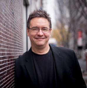 image of greg pliska. causasian man leaning against a brick wall with short salt and pepper (light brown) hair that's partially spiky. He's wearing wire-rim glasses, clean shaven with a nice smile. He is wearing a black t-shirt with a black blazer.
