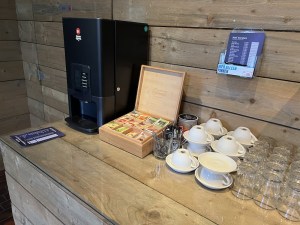 A coffee machine sits on a bar. Next to it is a selection of tag bags, coffee cups, and water glasses.