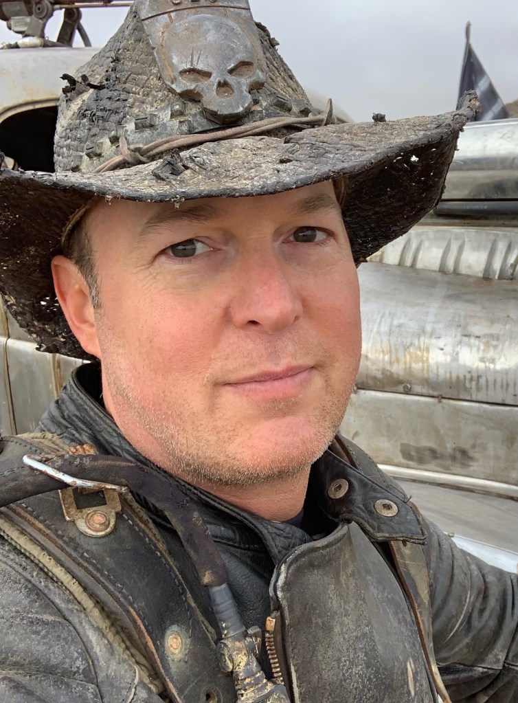 man wearing a dusty cowboy hat with a skull emblem and a dusty worn leather jacket.
