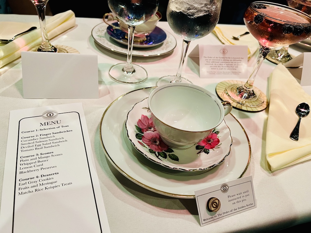 Closeup of a place setting for one at Order of the Golden Scribe. A teacup sits on a saucer, which sits on a plate. A pink mocktail glistens in the background.