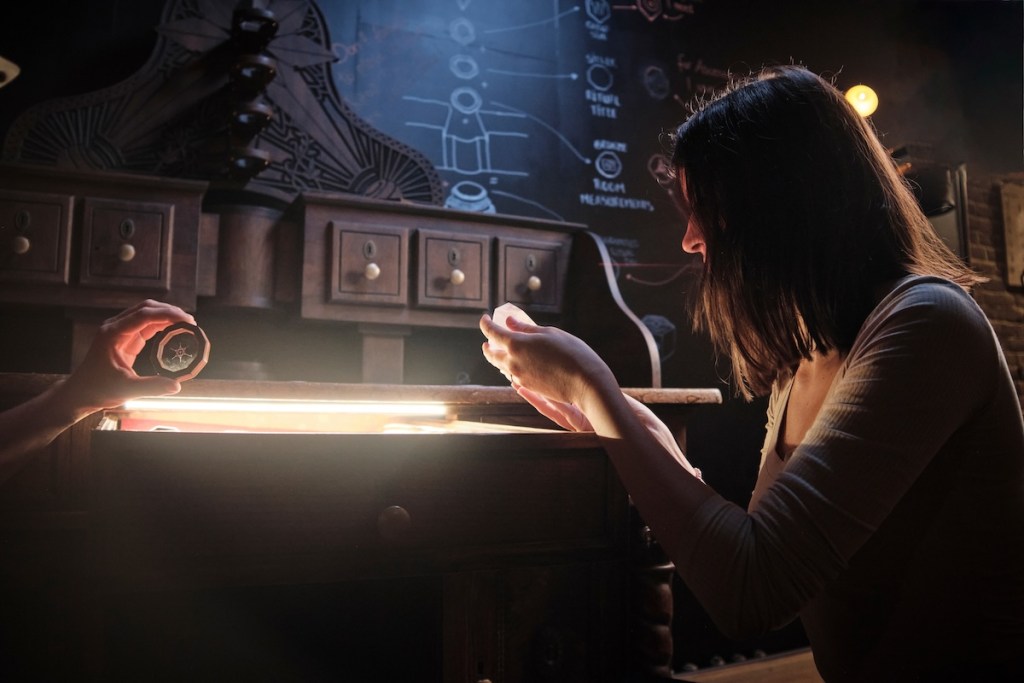 A women holds a small prop in her hand and looks at it intently. In the background is a wooden desk with lots of small drawers.