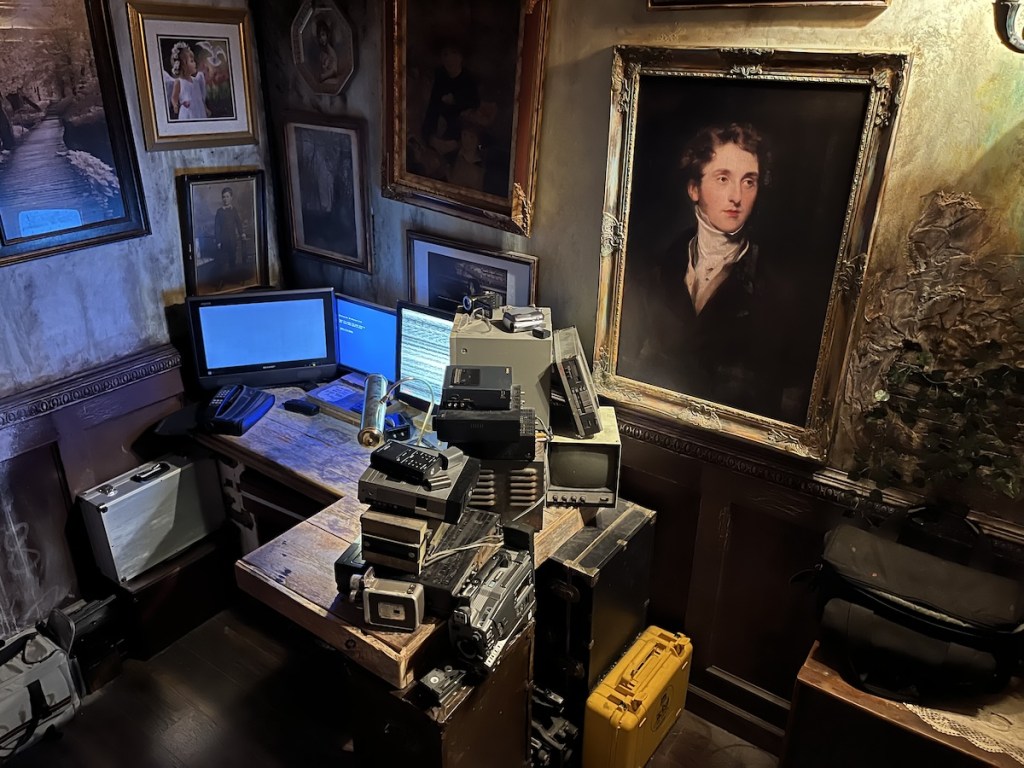 A room with framed paintings on the wall and desk covered in computers and other technology.