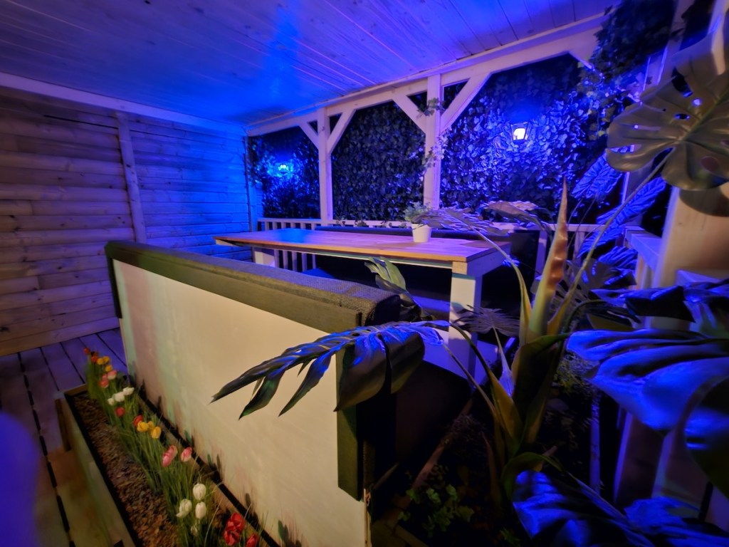 A table surrounded by benches sits on a wooden porch. There are plants and flowers in the foreground.