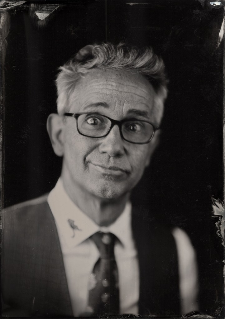black and white image of man in his 50's with salt and pepper hair, glasses and a wry smile, wearing a vest and tie with a button down shirt
