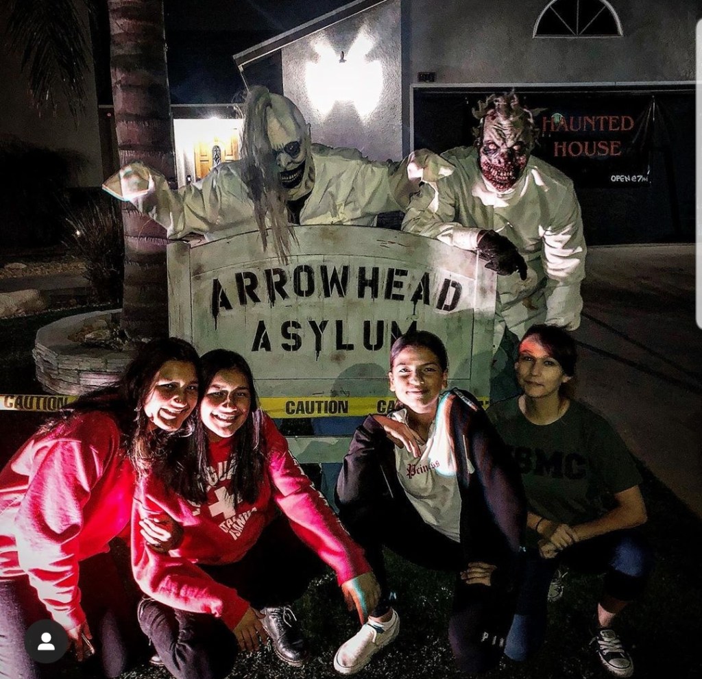 people at a home haunt posing in front of a sign that says arrowhead asylum. there are 2 actors in scary masks