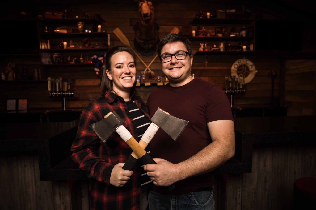 woman in plaid shirt with man in maroon t-shirt and glasses. they are holding crossed axes in front of a rustic bar.