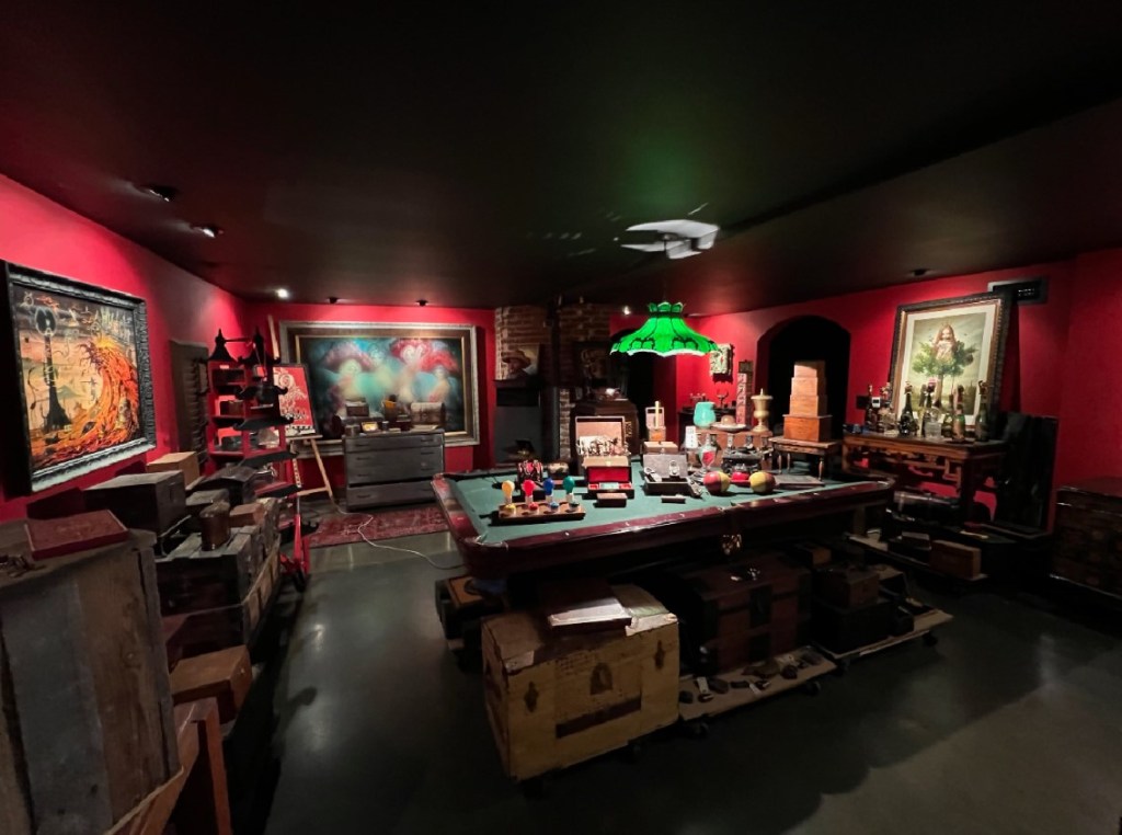 room with a pool table in the center. dimly lit with spot lights on groupings of boxes and collectible objects