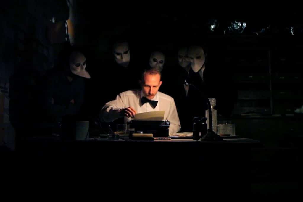 photo from sleep no more. man in white shirtsleeves and a bow tie is working on a typewriter. There are people surrounding him, wearing white masks. the room is shrouded in darkness