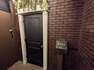 A wide-angle shot of an L-shaped cozy garden porch. Two plain black doors appear at the ends of the L. Each door has white framing, and plants dangle down from the ceiling and above each door. A string of large garden lights also hangs across the ceiling. In the middle of the L is a small brown shed.