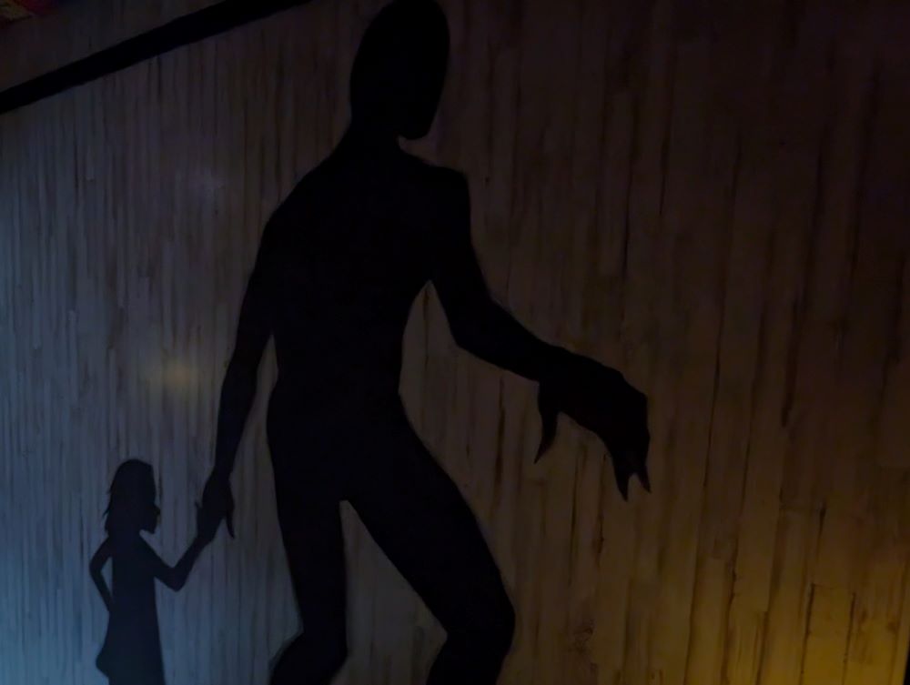 Two motionless, shadowy silhouettes appear walking to the right across a dimly lit, wooden-paneled wall. The leading shadow is a floor-to-ceiling giant. It is holding the hand of a small shadow child.