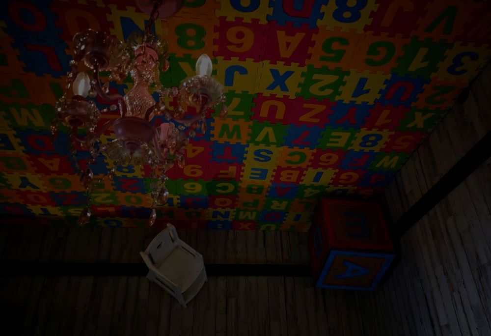 From the shadows, the brightly colored ceiling is barely visible. It is paved in alphabet tiles, and a pink crystal chandelier hangs to the left. A child's white rocking chair and large alphabet block protrude from the wall near the ceiling.