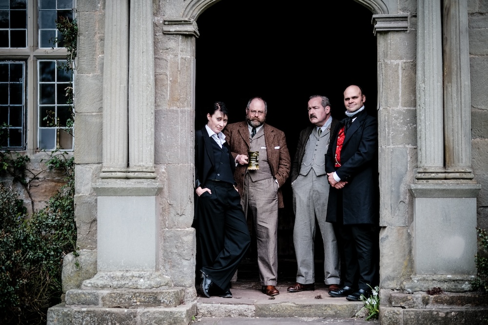 4 well-dressed performers, wearing suits of an earlier era, standing in an archway.