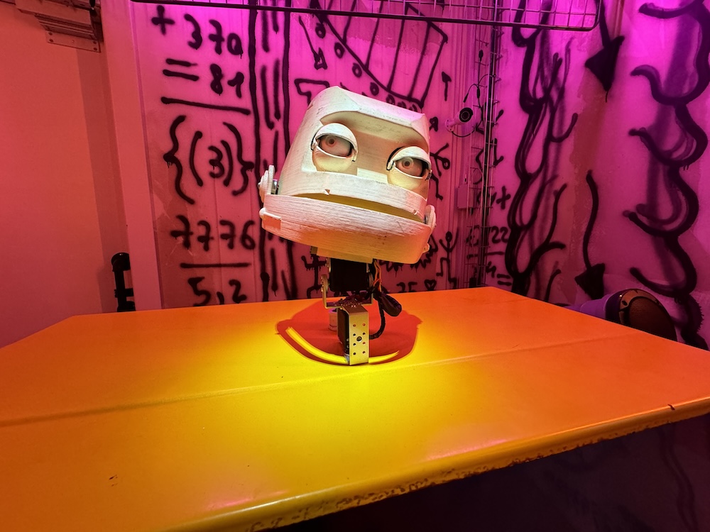 A small robotic character with large eyes on an orange stand.